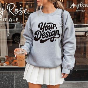 May include: A light gray sweatshirt with the text "Your Design" in black script. The person is wearing a white pleated skirt and holding a clear plastic cup with a straw. The background includes a storefront with pumpkins and fall decor.