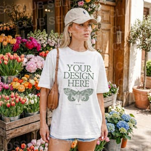 以下が含まれることがあります： 「YOUR DESIGN HERE」の緑色の文字と蝶のグラフィックが入った白いTシャツ。モデルはベージュの野球帽と茶色のショルダーバッグを着用しています。背景には花屋があります。