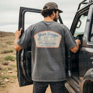 以下が含まれることがあります： 背面には「Your Design」と「Selfboss Mockup」の文字が入った、山と木の模様がデザインされたグレーのTシャツ。Tシャツは、ドアが開いた車の横に立っている人が着用しています。