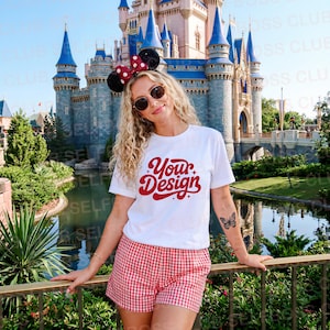 May include: A woman wearing a white t-shirt with the text "Your Design" in red script, paired with red and white checkered shorts. She is wearing sunglasses and a Minnie Mouse ear headband with a red bow and black ears. The background features a castle and a body of water.