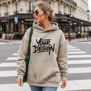 May include: A beige hoodie with a front pocket and a drawstring hood. The hoodie features the text "YOUR DESIGN" in black. The person in the image is wearing sunglasses and carrying a green bag.