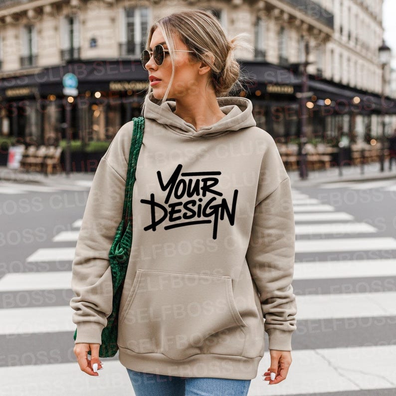 May include: A beige hoodie with a front pocket and a drawstring hood. The hoodie features the text "YOUR DESIGN" in black. The person in the image is wearing sunglasses and carrying a green bag.