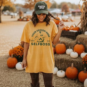 May include: A woman wearing a mustard yellow t-shirt and a green corduroy baseball cap with the text "PUMPKIN SPICE". The t-shirt has a graphic design with the text "YOUR DESIGN HERE", "ESTD 2020", and "SELFBOSS". Pumpkins and hay bales are in the background.