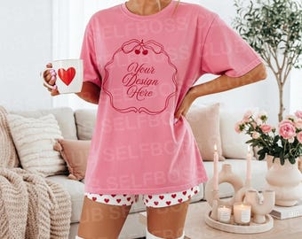 May include: A pink oversized t-shirt with the text "Your Design Here" inside a decorative frame, paired with white shorts featuring red hearts. The person is holding a white mug with a red heart. The outfit is completed with white knee-high socks.