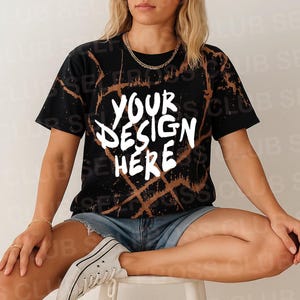 May include: A black and brown tie-dye t-shirt with the text "YOUR DESIGN HERE" in white. The shirt is being modeled by a person sitting on a white stool.