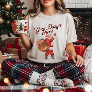 Può includere: Una maglietta color crema con una grafica di Babbo Natale e la scritta "Your Design Here". La persona tiene in mano una tazza natalizia con un manico rosso e un disegno di albero di Natale e Babbo Natale. Indossano pantaloni del pigiama a quadri rossi, verdi e bianchi e calzini fantasia.