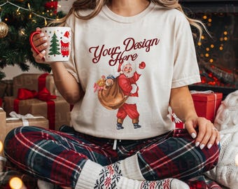 May include: A cream-colored t-shirt with a Santa Claus graphic and the text "Your Design Here." The person is holding a Christmas mug with a red handle and a Christmas tree and Santa Claus design. They are wearing red, green, and white plaid pajama pants and patterned socks.