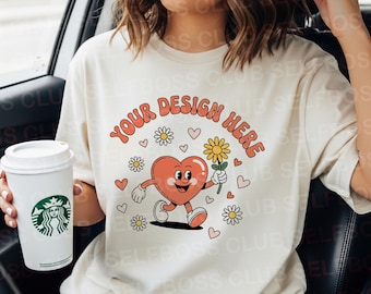 May include: A cream-coloured t-shirt with the text "YOUR DESIGN HERE" in orange. The shirt features a cartoon heart character holding a flower, surrounded by daisies and hearts. A Starbucks coffee cup is held in the left hand.