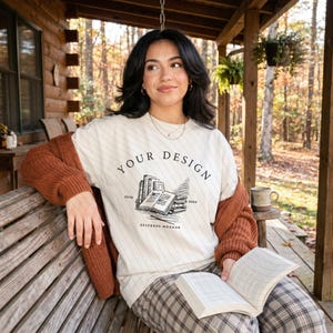 May include: A cream-colored t-shirt featuring a black line-art graphic of a stack of books and an open book. The text reads "YOUR DESIGN" in an arch above the books, with "ESTD 2020" on the sides and "SELFBOSS MOCKUP" printed below the books.