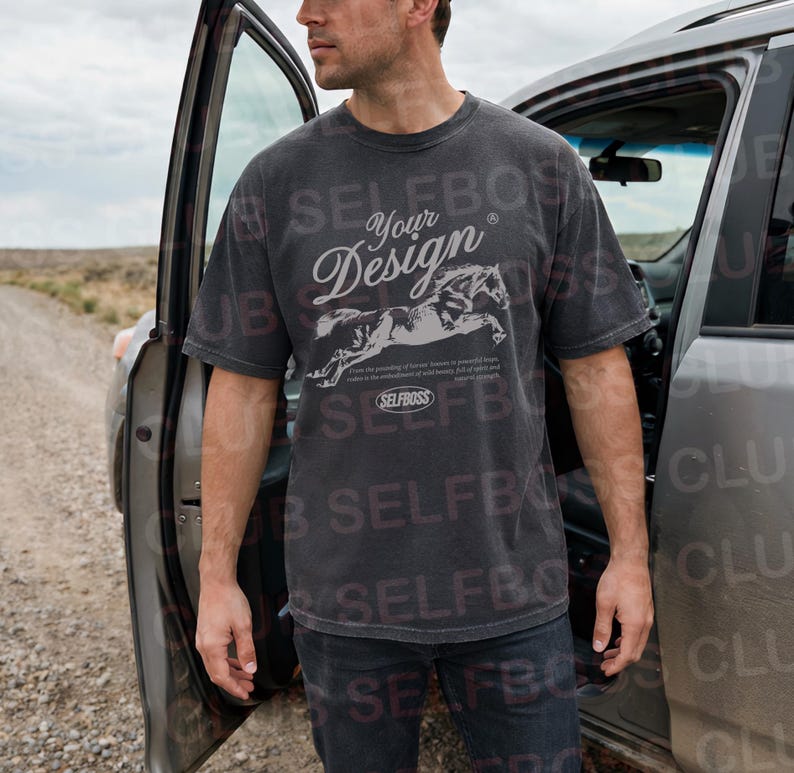 May include: A dark gray t-shirt with the text "Your Design" in a stylized font, along with a graphic of running horses. The shirt also features the text "SELFBOSS" and additional text below the horses. The shirt is worn by a person standing next to a car.