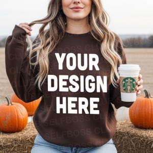 Może przedstawiać: Brązowa bluza z białym napisem "YOUR DESIGN HERE". Osoba trzyma biały kubek do kawy z zielonym logo. Dynie i bele siana w tle.