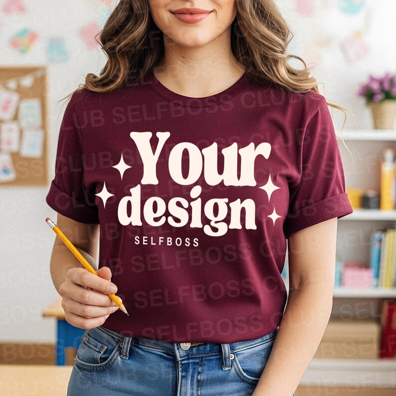 May include: Burgundy t-shirt with rolled sleeves featuring the text "Your design" in a retro font, with starburst accents. The word "SELFBOSS" is printed below the main text. The model is wearing blue jeans and holding a pencil.