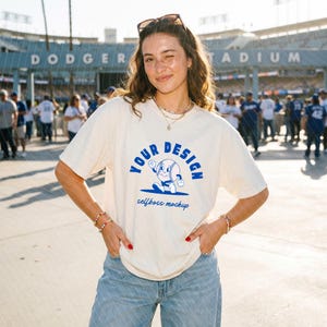 Op de afbeelding: Crèmekleurig T-shirt met een blauwe honkbal-thema grafiek met de tekst "YOUR DESIGN" en "selfboss mockup". Het shirt wordt gecombineerd met lichtblauwe jeans. De achtergrond toont een honkbalstadion.