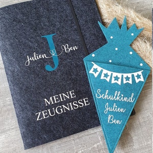 Puede incluir: Una carpeta de fieltro gris oscuro con una inscripci&oacute;n blanca que dice "Meine Zeugnisse" y una letra J de fieltro azul con los nombres "Julien & Ben" encima. Un cono de fieltro azul con estrellas blancas y el texto "Schulkind Julien Ben" est&aacute; a la derecha de la imagen.