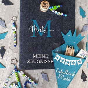 Puede incluir: Una carpeta de fieltro azul con el nombre "Mats" escrito en blanco. La carpeta tiene las palabras "Meine Zeugnisse" escritas en blanco. Hay un porta l&aacute;pices de fieltro azul con las palabras "Schulkind Mats" escritas en blanco. Tambi&eacute;n hay varias formas de fieltro azul y verde esparcidas alrededor de la carpeta.