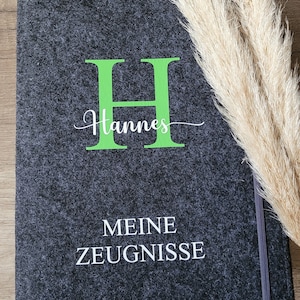 Puede incluir: Una carpeta de fieltro gris oscuro con una letra H verde y el nombre "Hannes" escrito en cursiva blanca. La carpeta tambi&eacute;n tiene las palabras "MEINE ZEUGNISSE" escritas en blanco.
