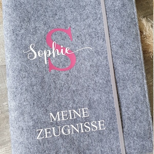 Puede incluir: Una carpeta de fieltro gris con una letra "S" rosa y el nombre "Sophie" escrito en cursiva blanca. La carpeta tambi&eacute;n tiene el texto "MEINE ZEUGNISSE" escrito en blanco.