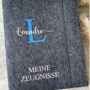 Puede incluir: Una carpeta de fieltro gris oscuro con una letra L azul y el nombre Leandro en letra cursiva. La carpeta tambi&eacute;n tiene el texto "MEINE ZEUGNISSE" en blanco.