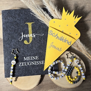 Puede incluir: Un cono escolar de fieltro amarillo con el texto "Schulkind Jonas" y una carpeta de fieltro negro con el texto "Jonas" y "Meine Zeugnisse". Hay dos collares de cuentas de alfabeto con un dije de abeja.