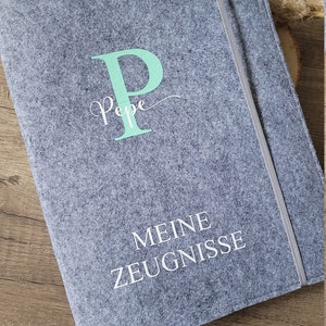 Puede incluir: Una carpeta de fieltro gris con una letra "P" verde claro y la palabra "Pepe" en letra cursiva. La carpeta tambi&eacute;n tiene el texto "MEINE ZEUGNISSE" en blanco.