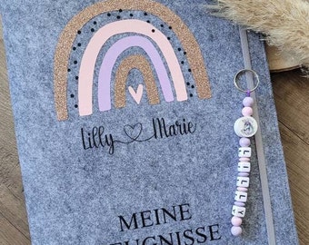 May include: Gray felt folder with a rainbow design, gold glitter accents, and the names "Lilly Marie" written in black. The folder has a zipper closure and a purple and pink beaded keychain with the name "Lilly Marie" on it. The text "MEINE ZEUGNISSE" is written in black at the bottom of the folder.
