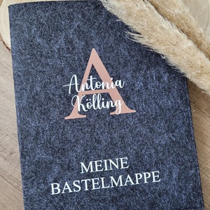 Puede incluir: Un libro de manualidades de fieltro gris oscuro con el texto "Antonia K&ouml;lling" y "MEINE BASTELMAPPE" en letras blancas. La letra "A" es una forma geom&eacute;trica grande de color oro rosa.