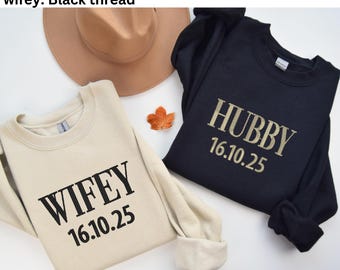 May include: Two sweatshirts, one beige and one navy blue, are displayed. The beige sweatshirt has the word "WIFEY" and the date "16.10.25" printed on it in black. The navy blue sweatshirt has the word "HUBBY" and the date "16.10.25" printed on it in beige.