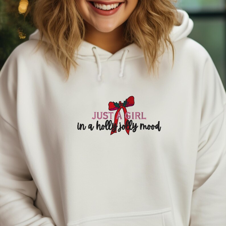 May include: White hoodie with a drawstring hood and a front pocket. The front of the hoodie features the text "JUST A GIRL in a holly jolly mood" with a red bow graphic.