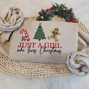 May include: Beige sweatshirt with the text "JUST A GIRL who loves Christmas" in red and black. The design includes a candy cane, Christmas tree, and gingerbread person. A beige scarf is draped around the sweatshirt.