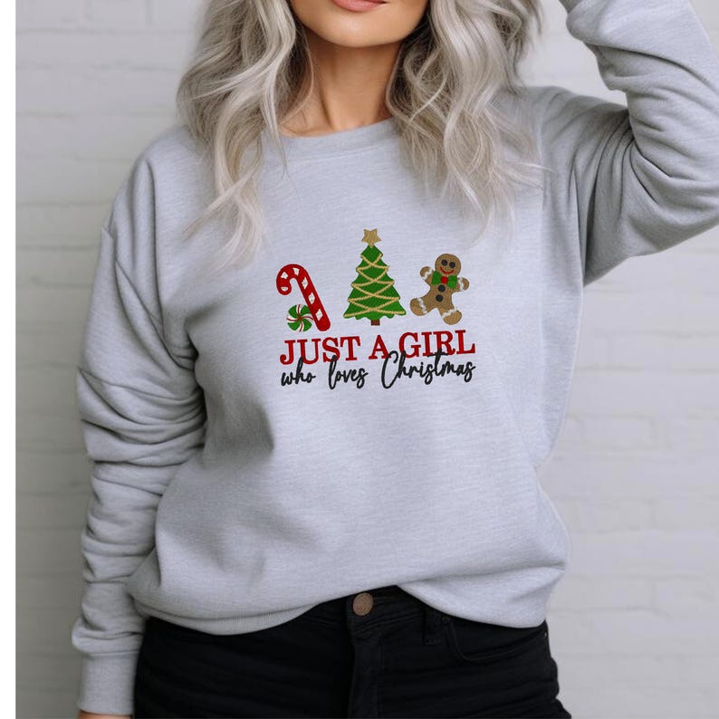 May include: A pale grey sweatshirt featuring a festive Christmas design. The design includes a red and green candy cane, a green Christmas tree, and a gingerbread man. The text reads "JUST A GIRL who loves Christmas."