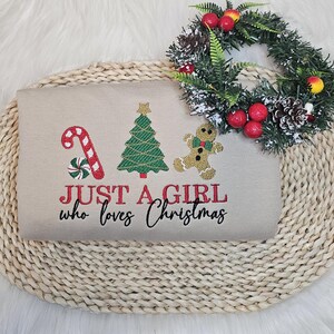 May include: A beige sweatshirt featuring embroidered Christmas motifs: a candy cane, Christmas tree, and gingerbread man. The text "JUST A GIRL who loves Christmas" is embroidered in red and black. A festive wreath with red berries and pinecones is placed above the sweatshirt.