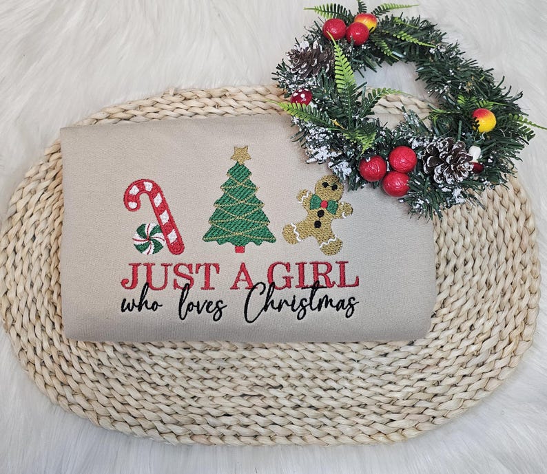 May include: A beige sweatshirt featuring embroidered Christmas motifs: a candy cane, Christmas tree, and gingerbread man. The text "JUST A GIRL who loves Christmas" is embroidered in red and black. A festive wreath with red berries and pinecones is placed above the sweatshirt.