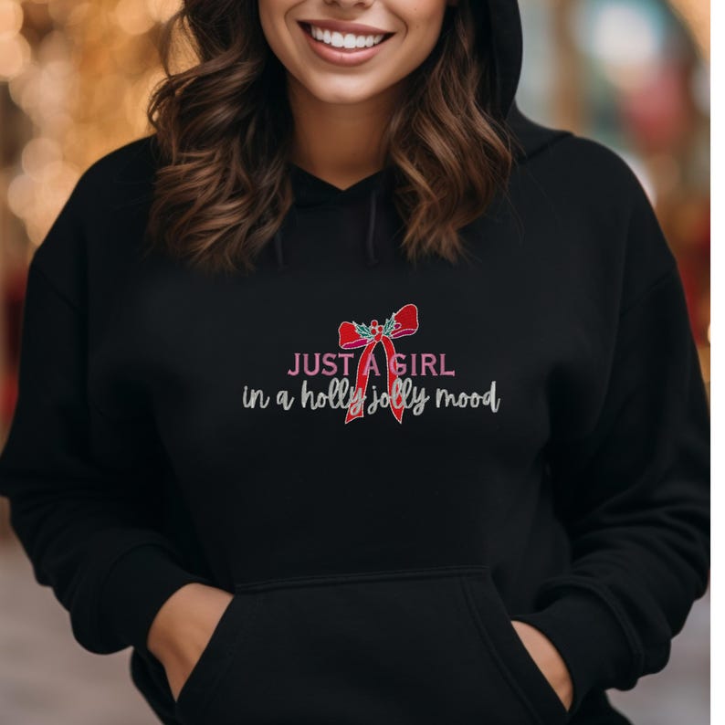 May include: Black hooded sweatshirt with a front pocket. The sweatshirt features the text "JUST A GIRL in a holly jolly mood" in white and pink, along with a red bow graphic. The design is centered on the chest.