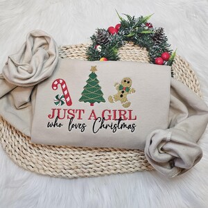 May include: Beige sweatshirt featuring Christmas embroidery: a candy cane, Christmas tree, and gingerbread man. The text "JUST A GIRL who loves Christmas" is in red and black. The sweatshirt is folded on a woven mat.