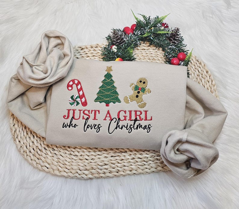 May include: Beige sweatshirt featuring Christmas embroidery: a candy cane, Christmas tree, and gingerbread man. The text "JUST A GIRL who loves Christmas" is in red and black. The sweatshirt is folded on a woven mat.