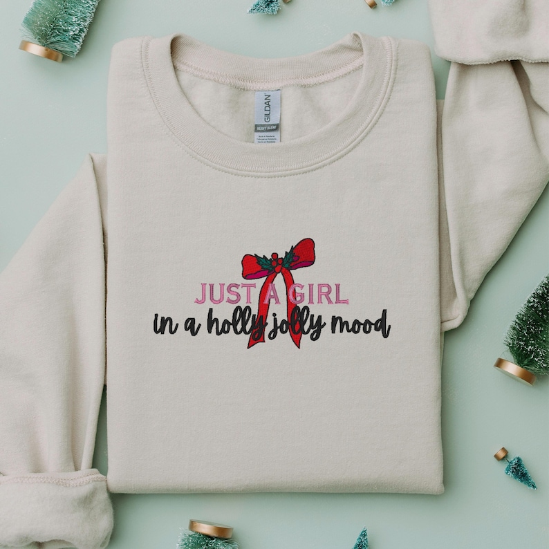May include: Cream-colored sweatshirt with the text "JUST A GIRL in a holly jolly mood" in pink and black script, accented by a red bow graphic. The sweatshirt is a Gildan brand.