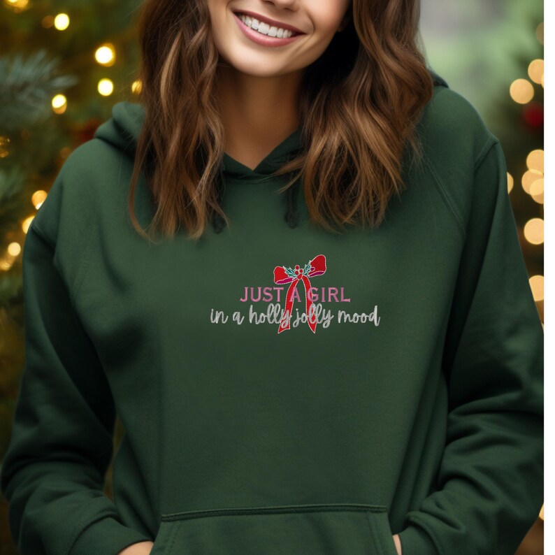 May include: Dark green hoodie with the text "JUST A GIRL in a holly jolly mood" in white and red. The design includes a red bow. The hoodie has a drawstring hood and a front pocket.