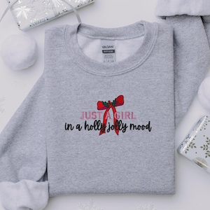 May include: A heather gray sweatshirt with the text "JUST A GIRL in a holly jolly mood" in pink and black, with a red bow graphic. The sweatshirt is a Gildan Heavy Blend.