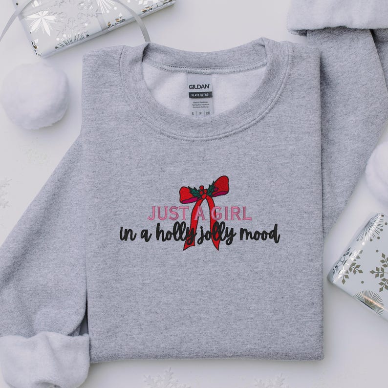 May include: A heather gray sweatshirt with the text "JUST A GIRL in a holly jolly mood" in pink and black, with a red bow graphic. The sweatshirt is a Gildan Heavy Blend.