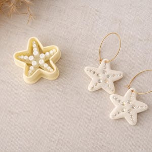 Op de afbeelding: Een stervormige koekjesvorm en twee paar oorbellen. De oorbellen en de koekjesvorm zijn wit en versierd met kleine, heldere strass-steentjes. De oorbellen hebben goudkleurige hoepels.