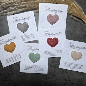 Könnte beinhalten: Fünf herzförmige Stoffaufnäher in Grau, Rosa, Rot, Senfgelb und Salbeigrün, befestigt auf weißen Karten mit dem Text Kleiner Alltagsbegleiter und einem deutschen Gedicht. Die Aufnäher haben eine strukturierte, gewebte Oberfläche.