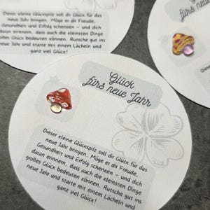 Könnte beinhalten: Runde weiße Karten mit einer kleinen Glaspilz-Figur in Rot, Gelb oder Blau. Jede Karte zeigt eine vierblättrige Kleeblatt-Illustration und den deutschen Text Glück fürs neue Jahr, gefolgt von einem Absatz mit Wünschen für Freude, Gesundheit und Erfolg.