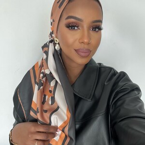 May include: A woman wearing a brown and black patterned headscarf with a black leather jacket. She is wearing gold earrings and has a nose ring.