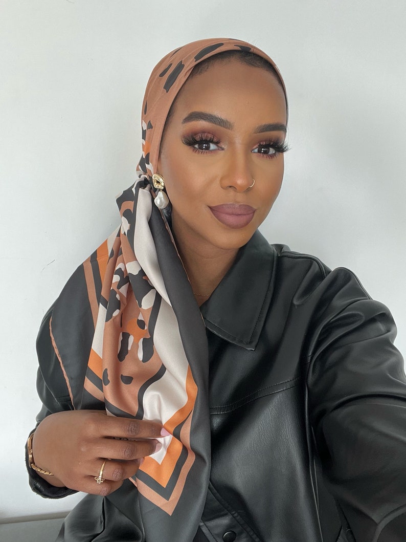 May include: A woman wearing a brown and black patterned headscarf with a black leather jacket. She is wearing gold earrings and has a nose ring.