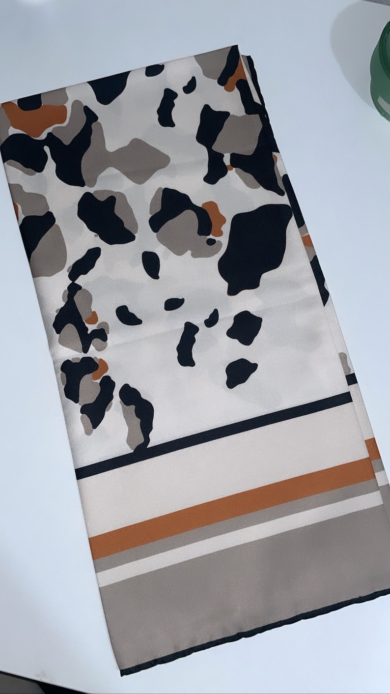 May include: A white scarf with a black, brown, and orange leopard print pattern. The scarf has a black border and a brown and white striped border.