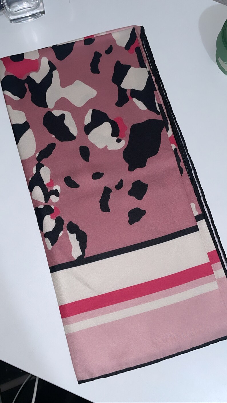 May include: A pink and black patterned scarf with a white and pink striped border. The scarf has a black trim around the edges.