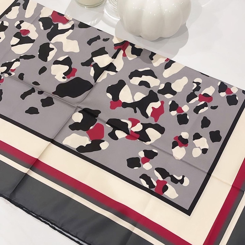 May include: A gray silk scarf with a leopard print design in black, white, and red. The scarf has a cream border with a red and gray stripe.
