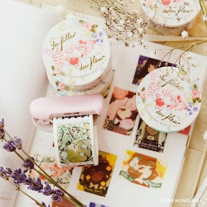 Puede incluir: Una colección de rollos de cinta adhesiva decorativa y un dispensador de cinta rosa. Los rollos de cinta adhesiva presentan ilustraciones florales y de chicas con el texto "Les filles des fleur". El dispensador contiene un rollo de cinta con el texto "Muguet".