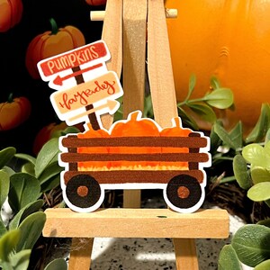 May include: A wooden easel with a pumpkin-themed sticker. The sticker features a red wagon filled with orange pumpkins and two signs that read "Pumpkins" and "Hay Ride".
