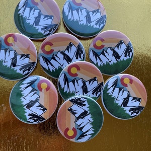 May include: A collection of round Colorado-themed buttons. Each button features a mountain range silhouette, a sunset gradient, and the Colorado state flag. The buttons are in shades of green, orange, yellow, and blue.
